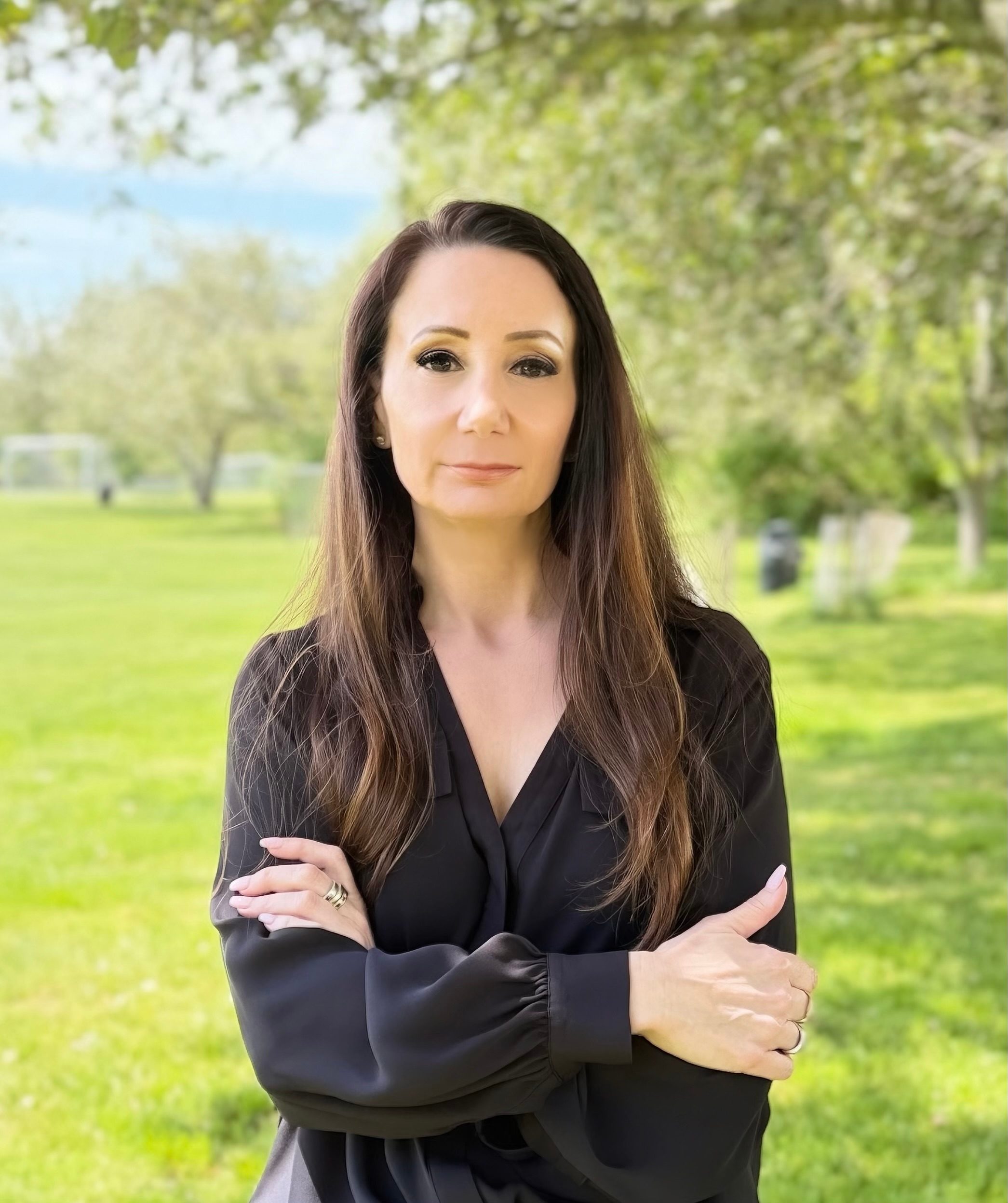 Woman business owner with long hair wears black blouse and smiles at camera with crossed arms while standing in a grassy park