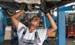 A young African American female mechanic is standing under a car and examining the damage on the broken parts with a serious look on her face.