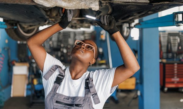 A young African American female mechanic is standing under a car and examining the damage on the broken parts with a serious look on her face.