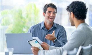 Two white men discuss finances in front of laptop and tablet